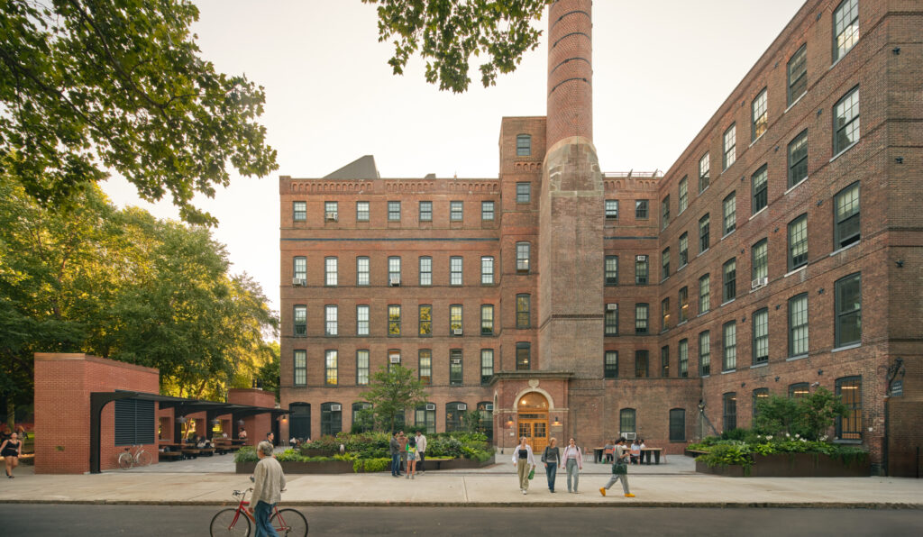 The image depicts an outdoor scene at Pratt Institute, showcasing a historic brick building with large, multi-pane windows and a tall chimney. The building is surrounded by lush greenery. In the foreground, a few people are engaged in casual activities, such as walking and conversing, near a modern brick structure that appears to be a bike storage area. The setting conveys a vibrant, academic environment bathed in the warm, soft light of a late afternoon.