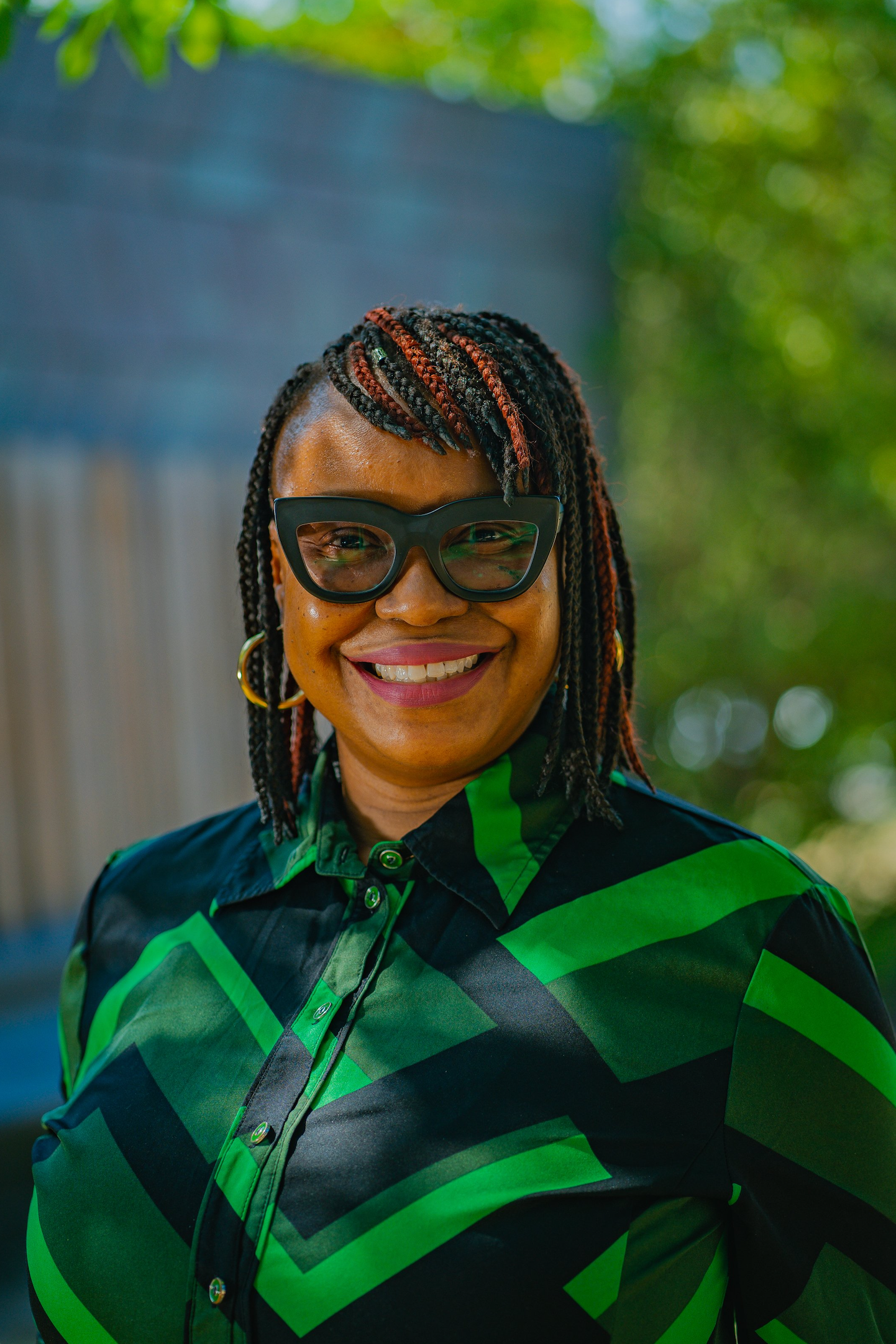 A woman wearing oversized black glasses and a green and black geometric-patterned blouse is smiling in an outdoor setting with a blurred background of green foliage.