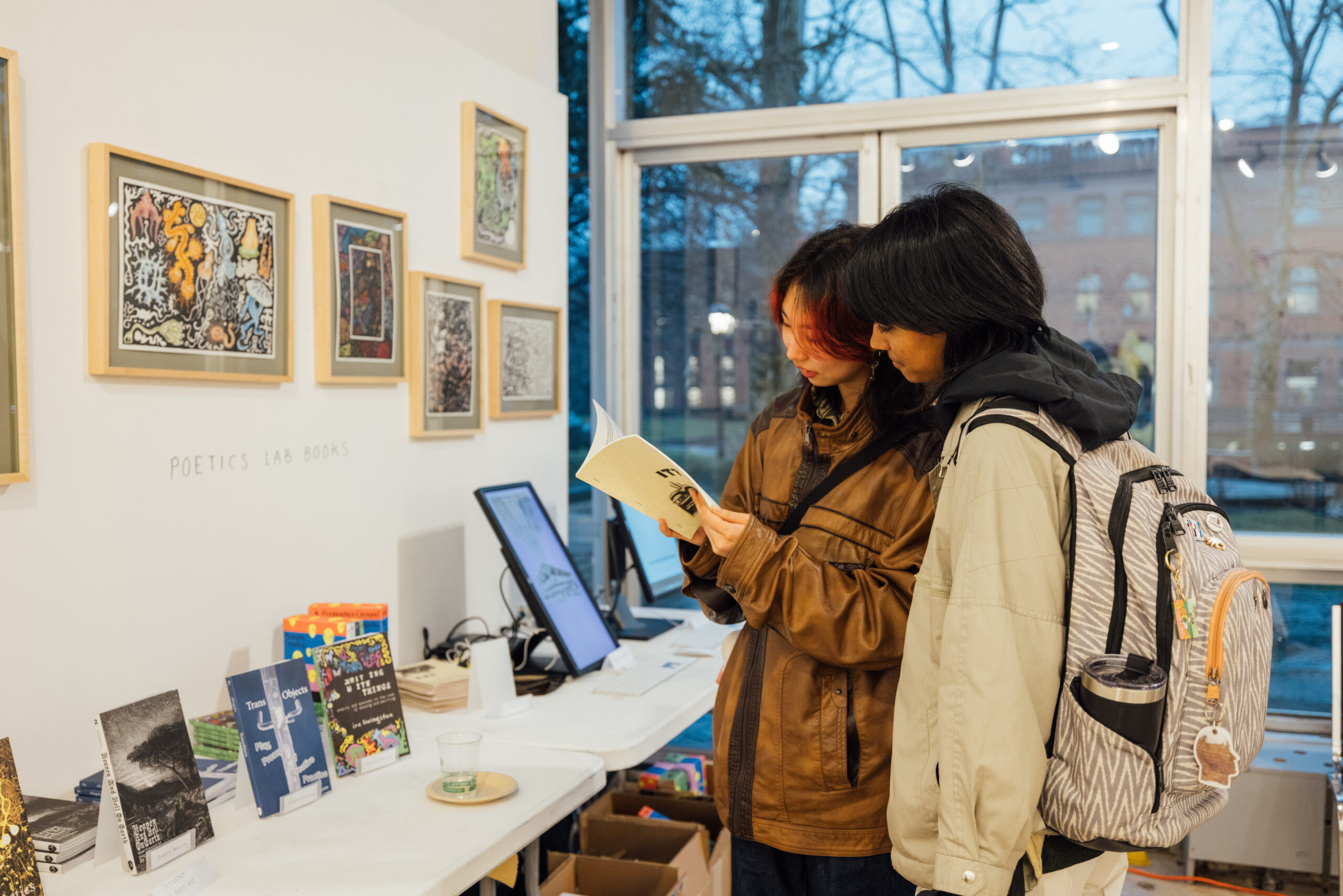 Two students look at a small book together side-by-side in a room across from two tables pushed against a wall featuring framed prints