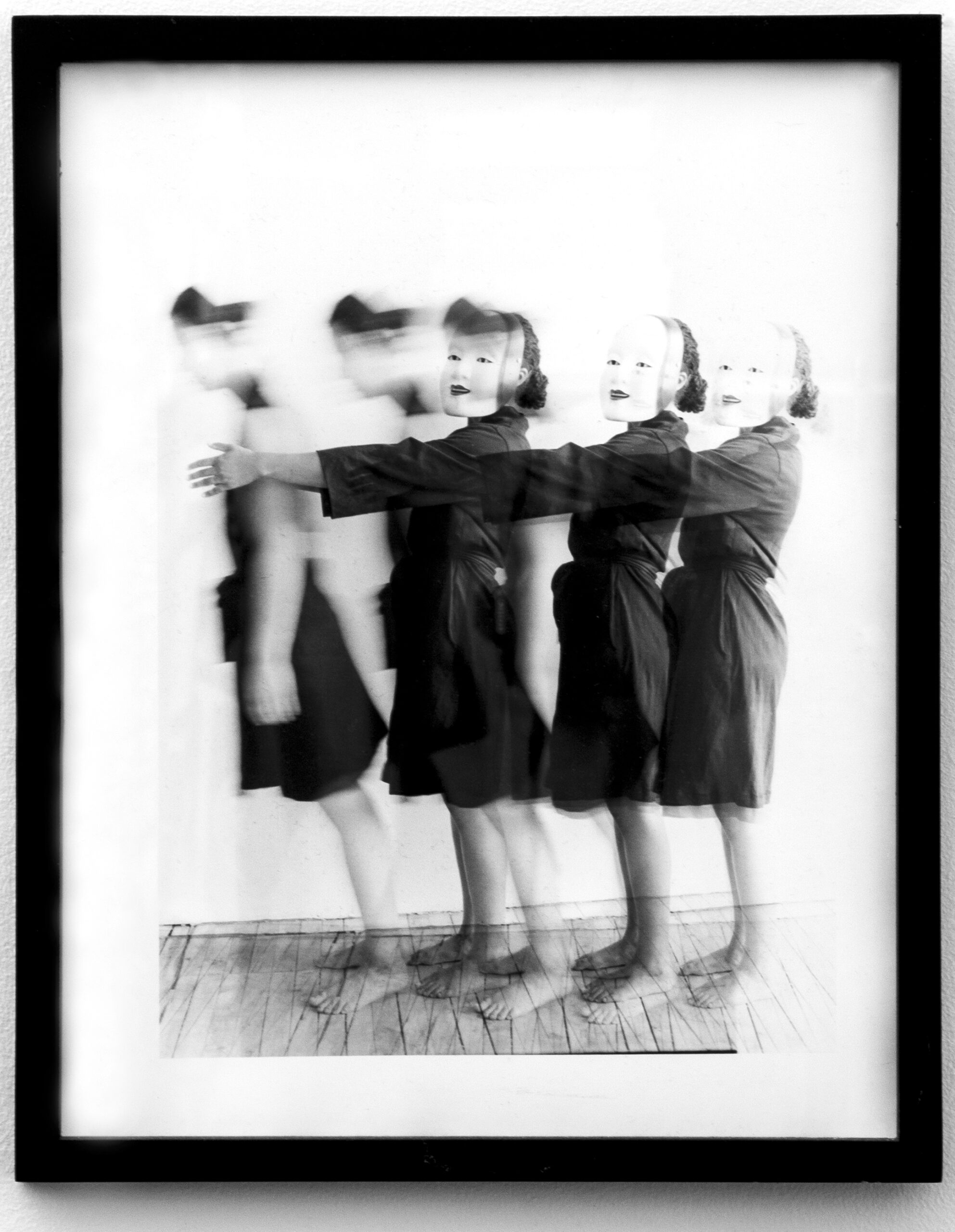 A black and white photograph showing a blurred motion effect of a figure in a dress, captured multiple times in sequence. The figure appears to be in mid-movement, creating a ghostly, layered visual effect. The individual wears a mask, adding to the surreal atmosphere. The image is framed with a black border.