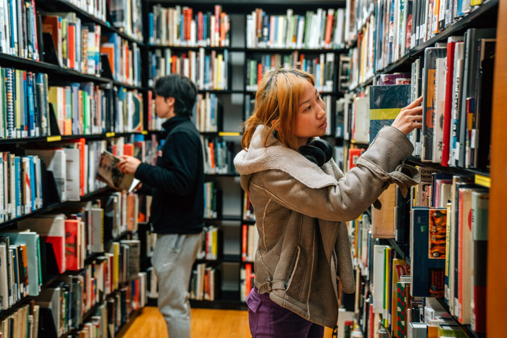 Two individuals browsing books in a library. A woman in the foreground, wearing a tan shearling jacket and headphones around her neck, examines a book on the right shelf. In the background, a man in dark clothing reads a book on the left shelf. The shelves are filled with various colorful books.