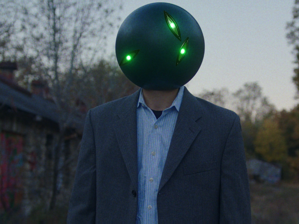 A surreal scene of a person wearing a formal suit jacket and collared shirt, standing outdoors in front of an abandoned building and trees. Instead of a human head, the person has a dark spherical head with three glowing green eye-like shapes, creating an otherworldly, science fiction-inspired effect.