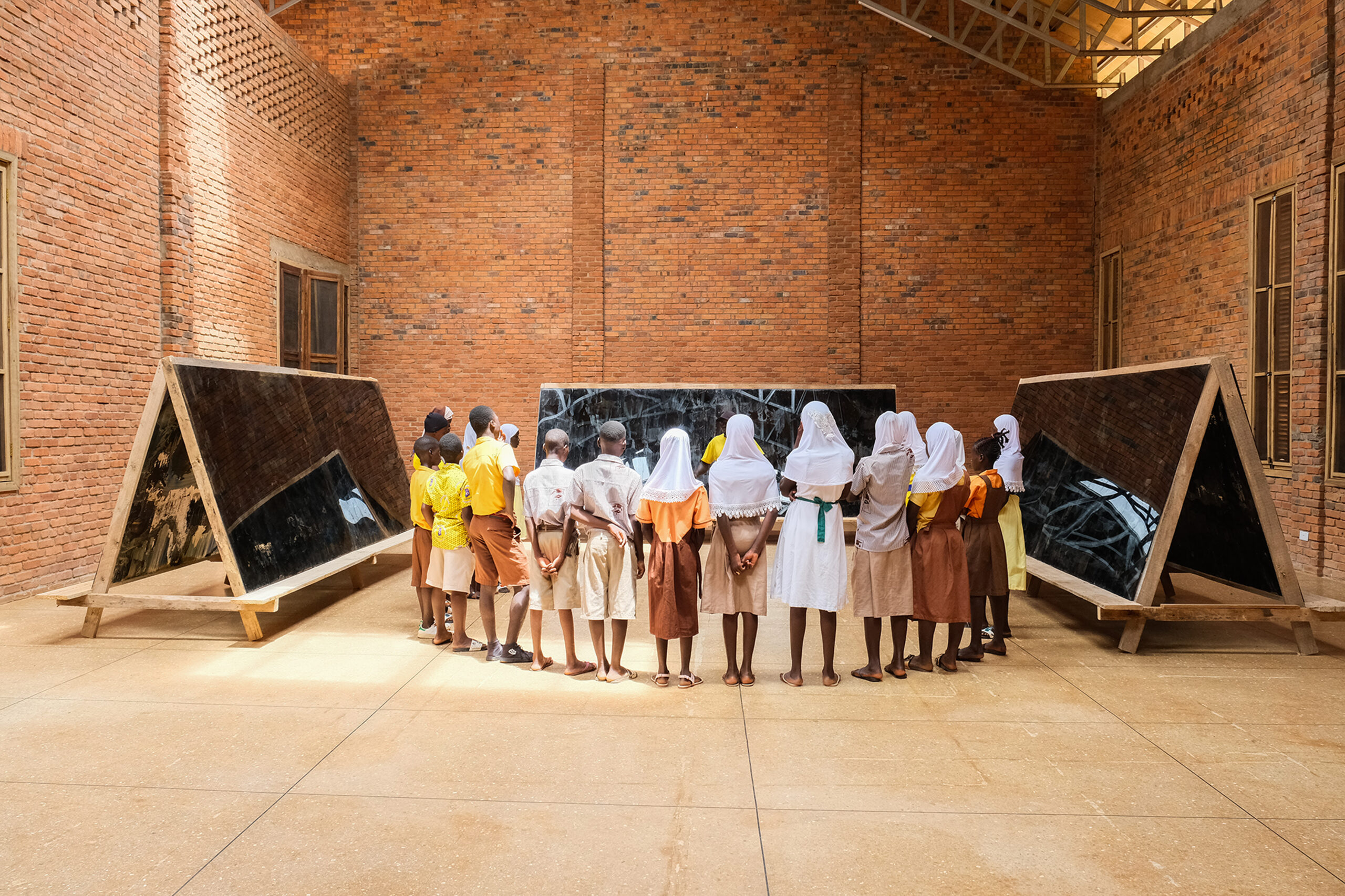 A group of children in school uniforms stand in two rows, observing large triangular mirror-like structures in an open indoor space with brick walls. The children are mostly wearing light brown and yellow clothing, with some wearing headscarves. The room has high ceilings and natural light.