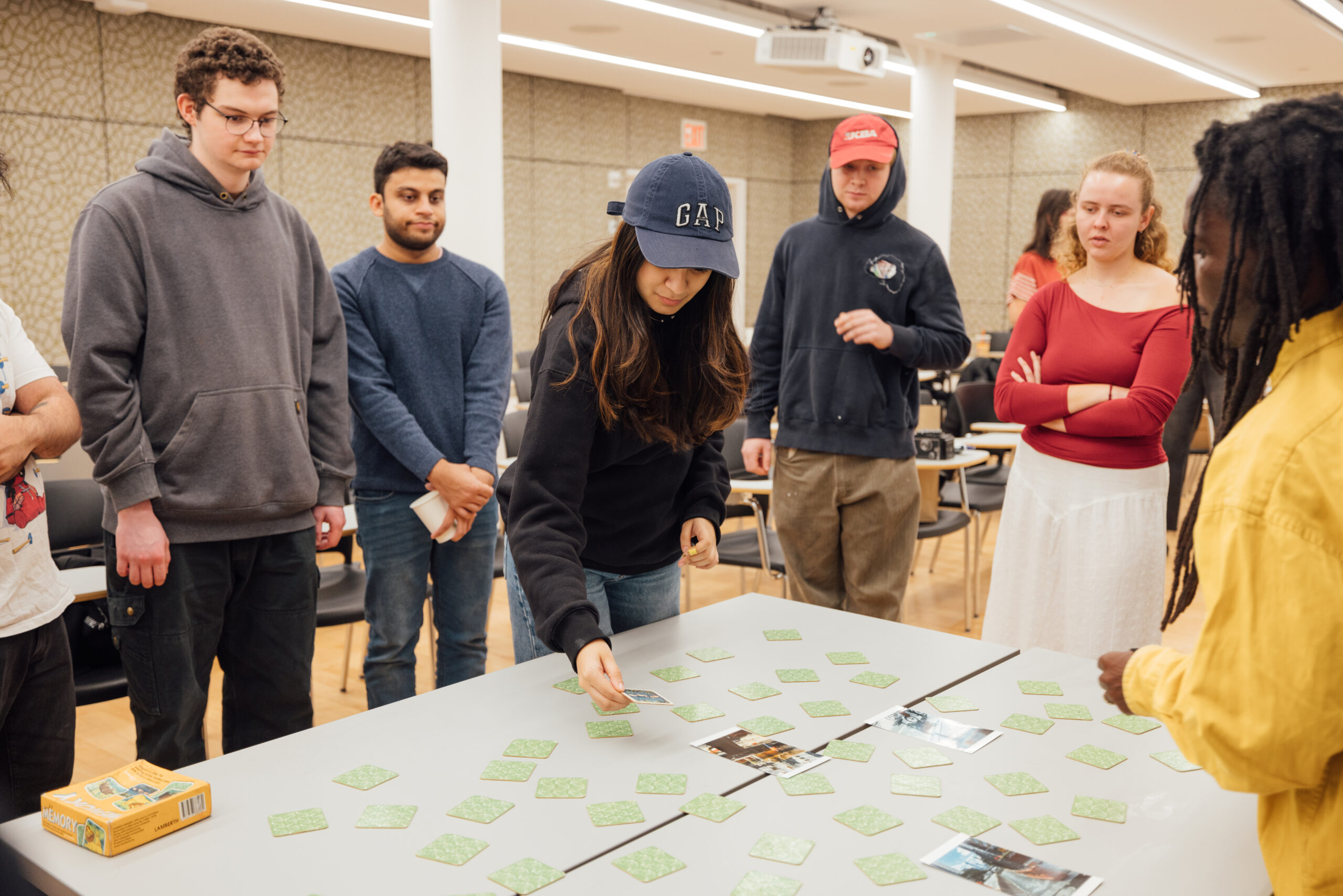 A group of students gathered around a table during a classroom activity. One student wearing a navy blue 'GAP' hat is selecting a card from a grid of green-backed cards spread across the table, while others watch attentively. The setting is a brightly lit classroom with chairs and a projector visible in the background.