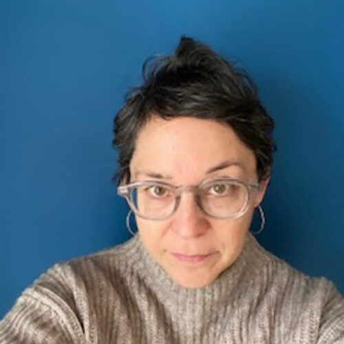 A person with short dark hair and glasses, wearing a gray sweater, looking directly at the camera. The background is a solid blue wall.