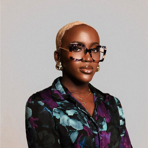 A portrait of a person with short blonde hair, wearing large tortoiseshell glasses and a floral top, looking confidently at the camera. The background is neutral gray.