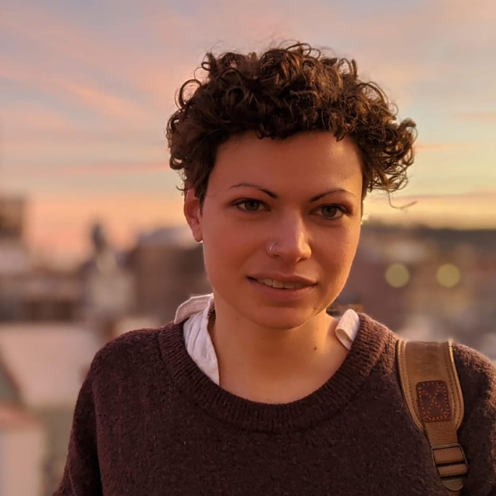 A person with short curly hair and a nose ring, wearing a dark sweater and standing outdoors during a sunset. The background features soft pink and orange hues.