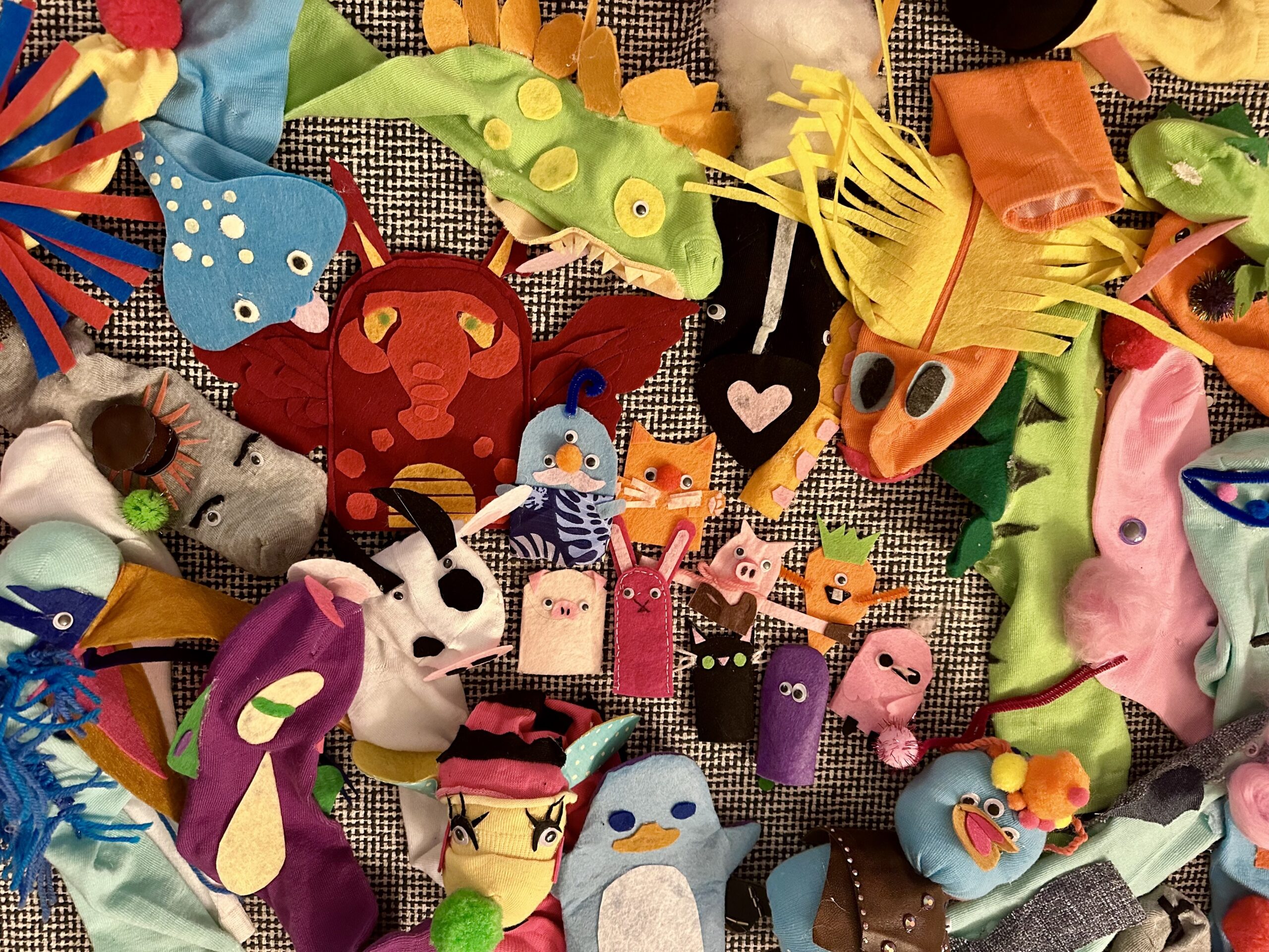 A collection of handmade felt puppets on a table. The puppets are crafted with vibrant colors and various materials, depicting animals, abstract shapes, and other characters.