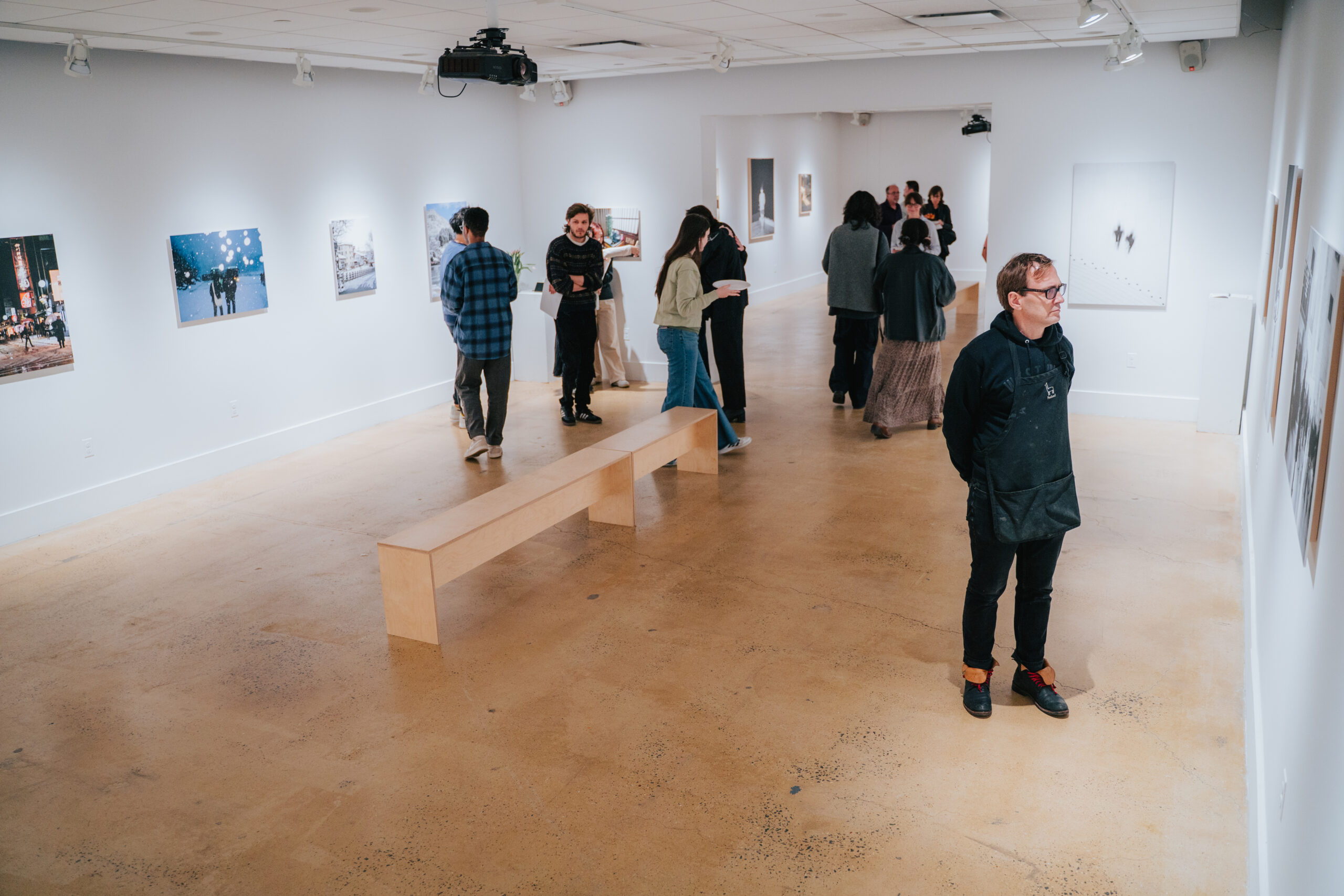 A woman stands observing a framed photograph of a snowy scene on the wall. Two other visitors are in the background, examining different pieces. The gallery has clean white walls, with bright overhead lighting and minimal decor.