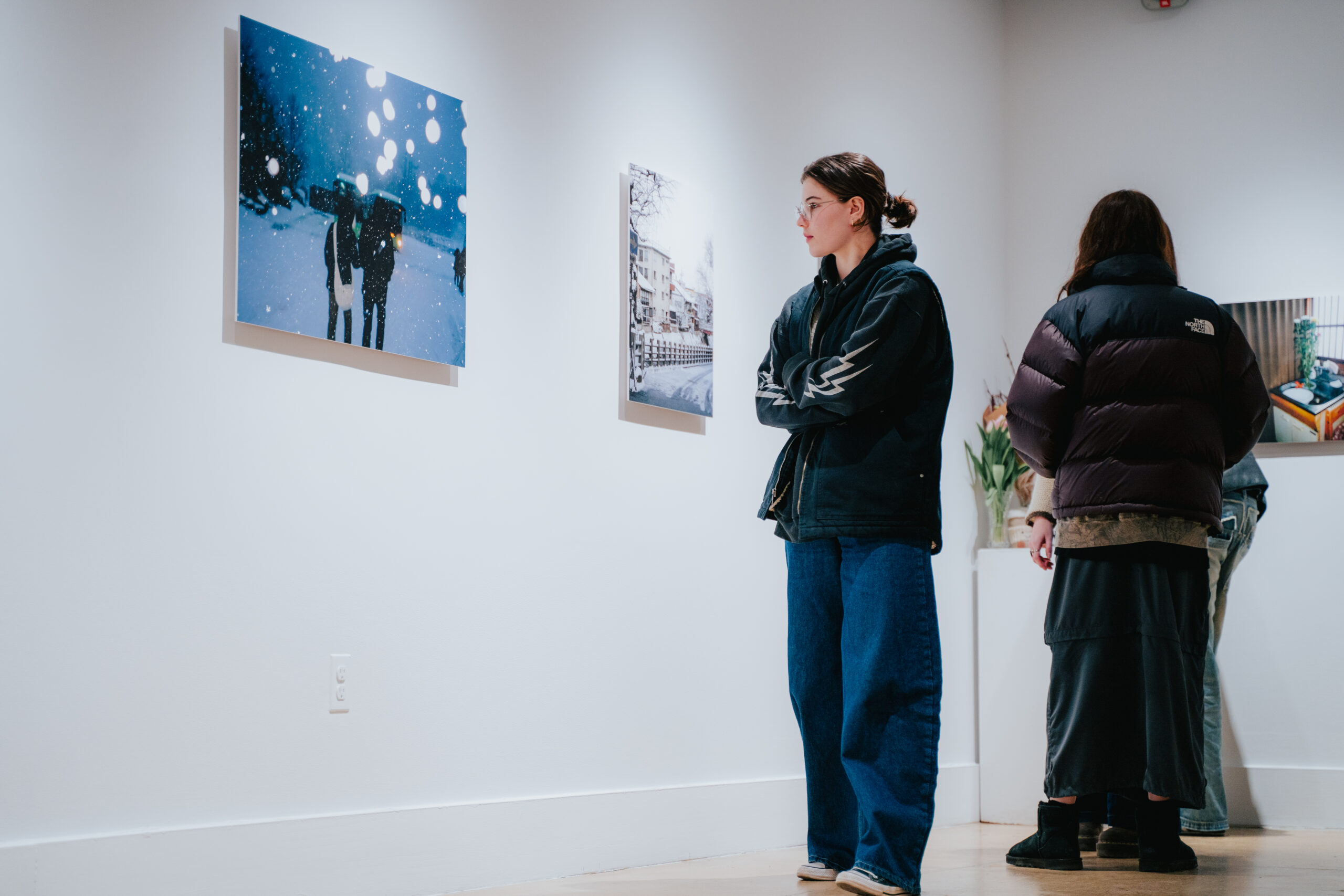 Gallery visitors are viewing artwork on white walls. Two individuals are seated on a wooden bench to the left, while others stand closer to the framed photographs on display. The ceiling features recessed lighting, and the gallery floor is polished concrete.