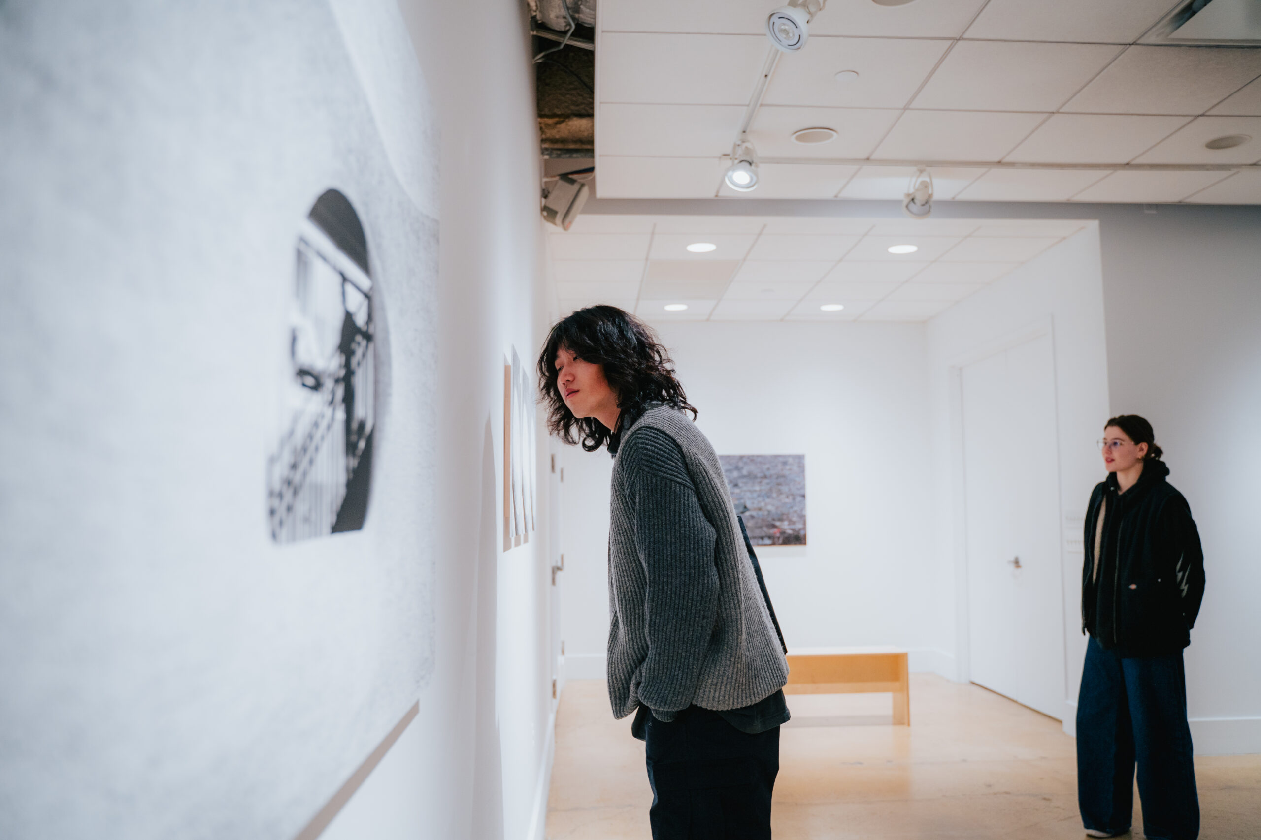 A young man walks along a gallery wall lined with framed photographs, hands clasped behind his back. The artworks are mounted evenly on a white wall under focused lighting, with polished concrete floors adding to the clean aesthetic.