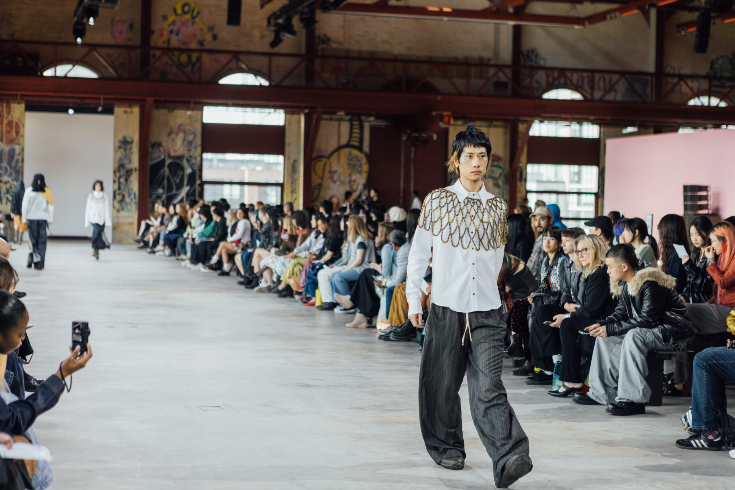 A runway fashion show taking place in a spacious industrial-style venue with graffiti-decorated walls. A model walks confidently down the runway wearing a white shirt with a gold chain design and loose-fitting dark trousers. The audience is seated along both sides of the runway, many capturing the moment on their phones. The atmosphere is vibrant and creative, with natural light streaming through large windows.