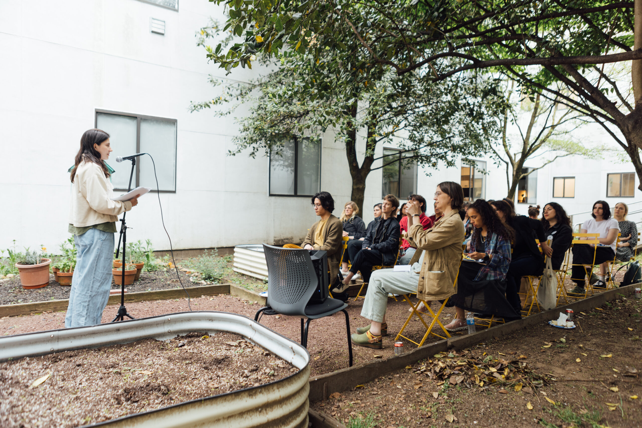 A woman stands at a microphone reading from papers in an outdoor garden setting. Behind her, a small audience of people sits on yellow chairs, attentively listening. The scene is surrounded by trees, plants, and white building walls in the background, creating a casual and serene atmosphere.