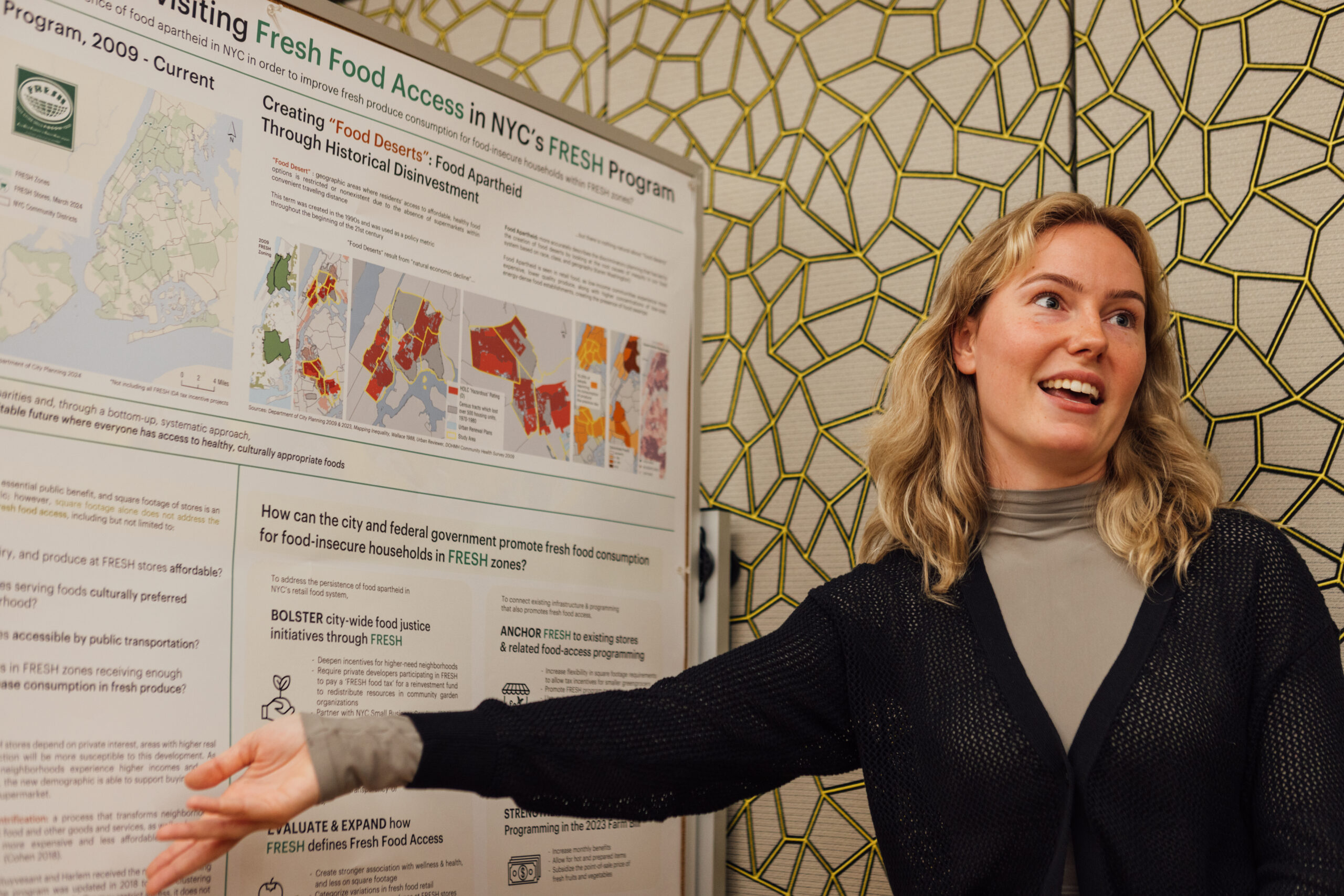 Woman standing at presentation board, wearing black cardigan sweater over pink t-shirt, with blond hair, smiling. The woman points at information for 