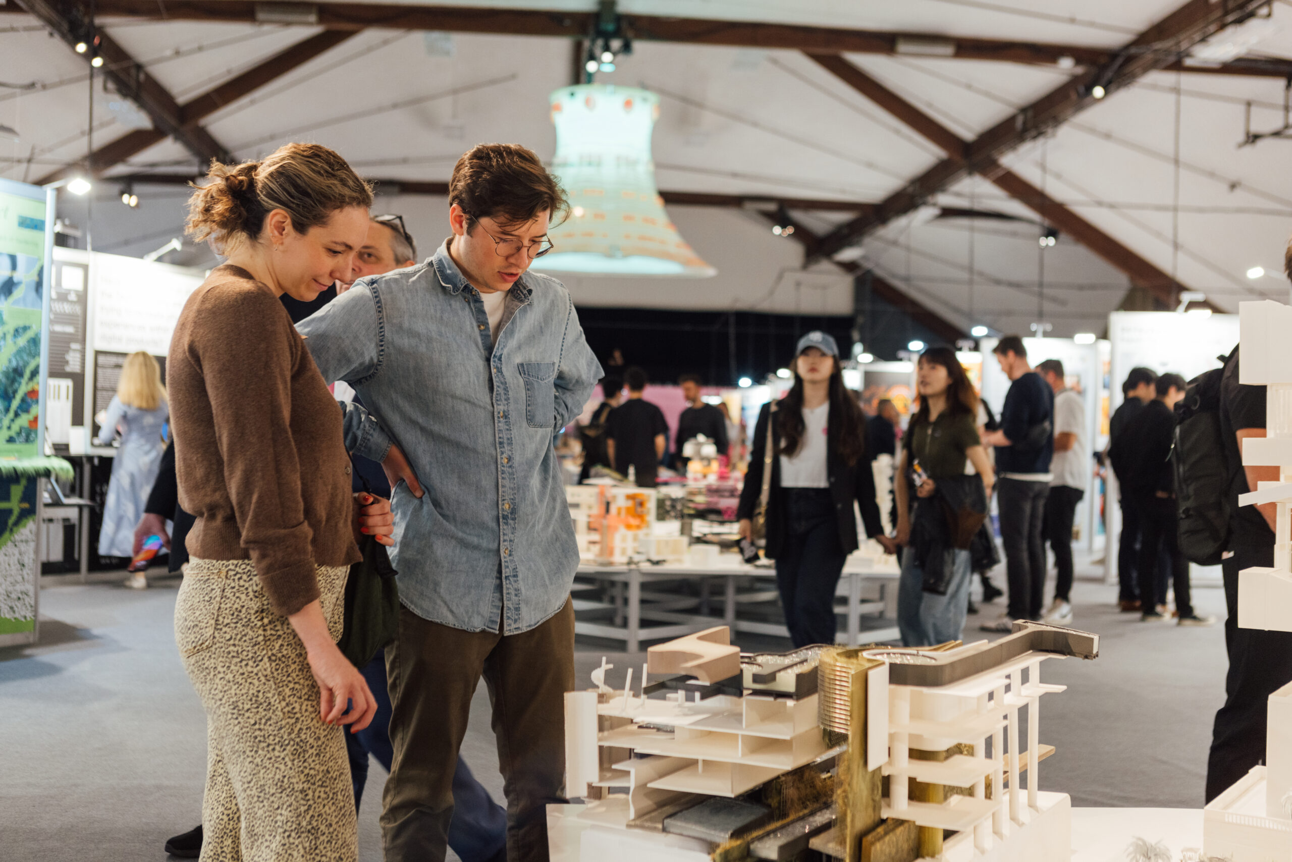 Two individuals, a man and a woman, closely examine an architectural model displayed in an indoor exhibition space. Other attendees are visible in the background, walking and observing various exhibits. The venue features high ceilings with wooden beams and bright, focused lighting, creating an engaging and professional atmosphere.