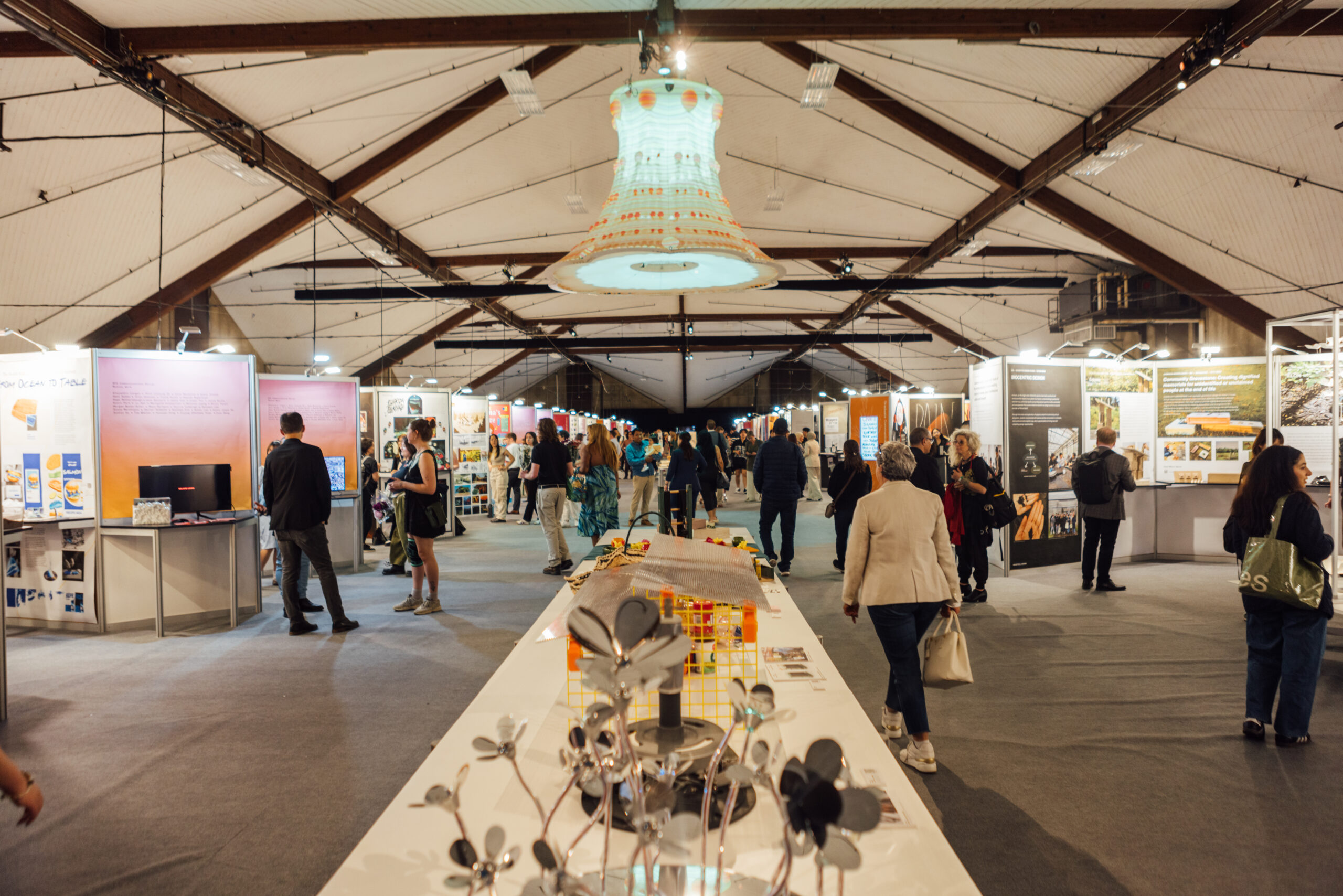 A wide-angle shot of a exhibition hall, with a large centerpiece installation in the middle of the walkway, with booths on each side of the hall, with artworks on display.