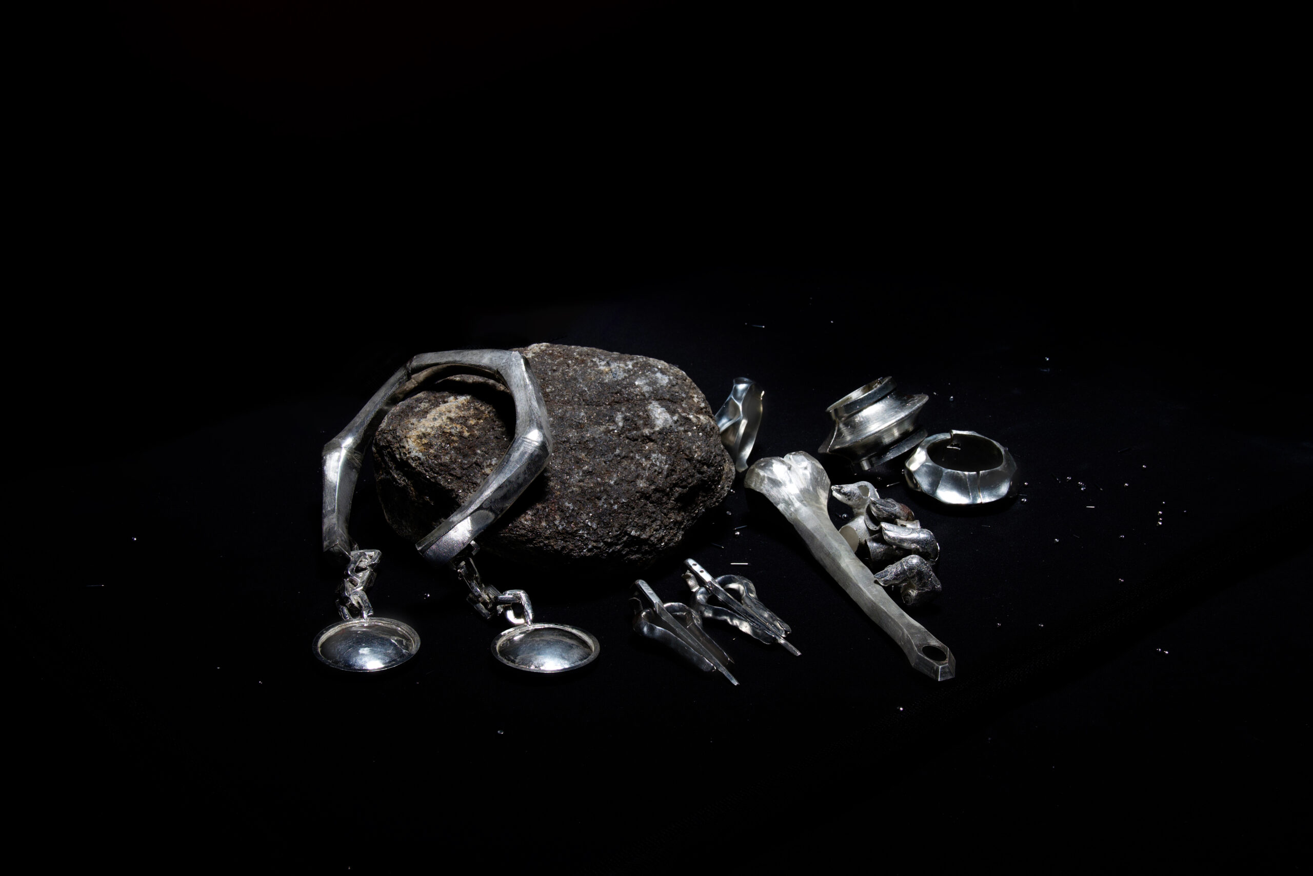 A collection of handcrafted silver jewelry pieces arranged on a black surface alongside a large textured rock. The jewelry includes bracelets, rings, and other sculptural designs, highlighted against the dark background to emphasize the metallic sheen and intricate craftsmanship.