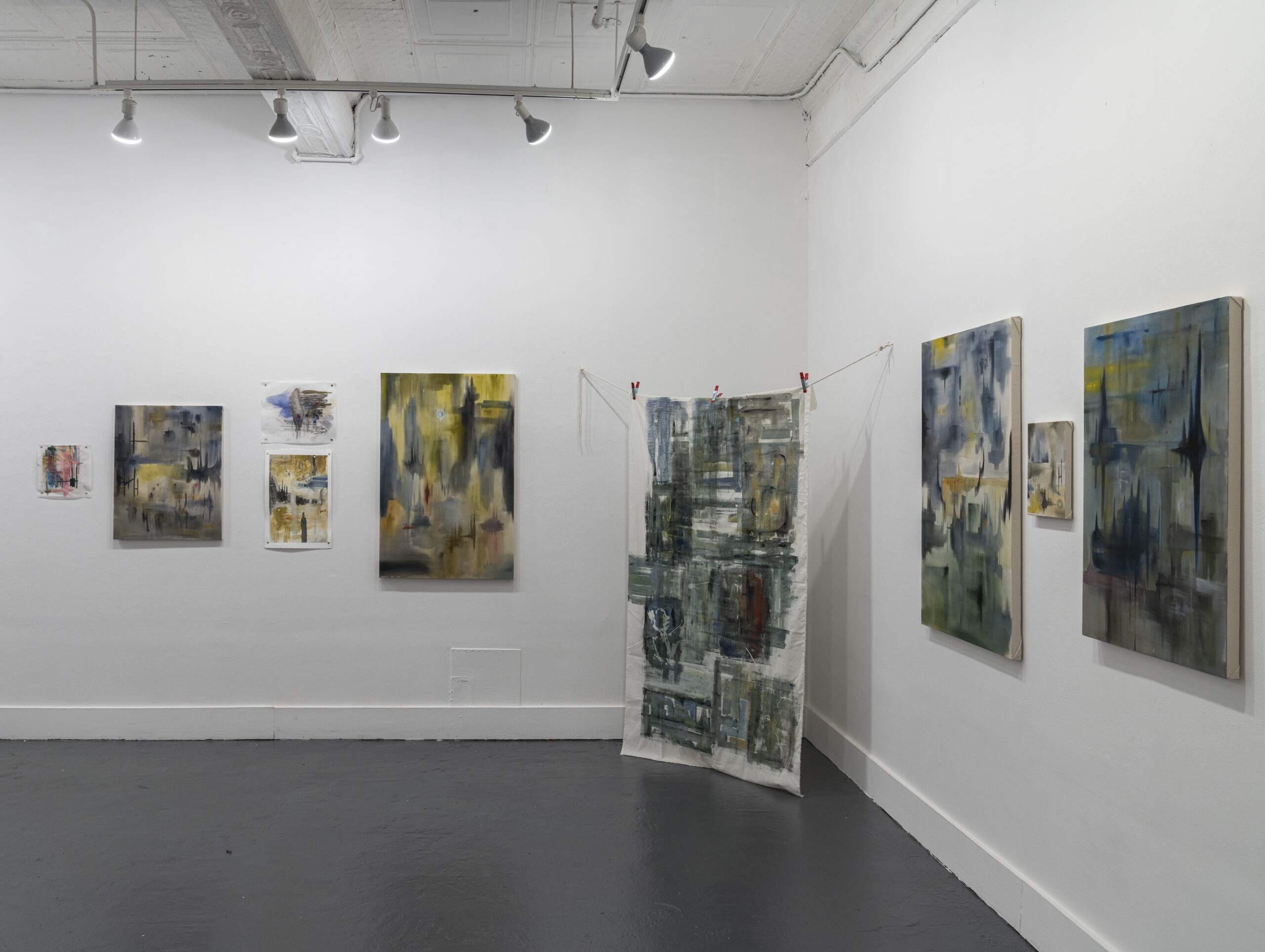 A gallery installation showcasing abstract paintings in subdued tones of blue, yellow, and gray. Several canvases hang on a white wall, with one fabric artwork displayed in the center, clipped onto a string. The room is minimally lit with spotlights, emphasizing the artworks' texture and depth against a clean, contemporary space.