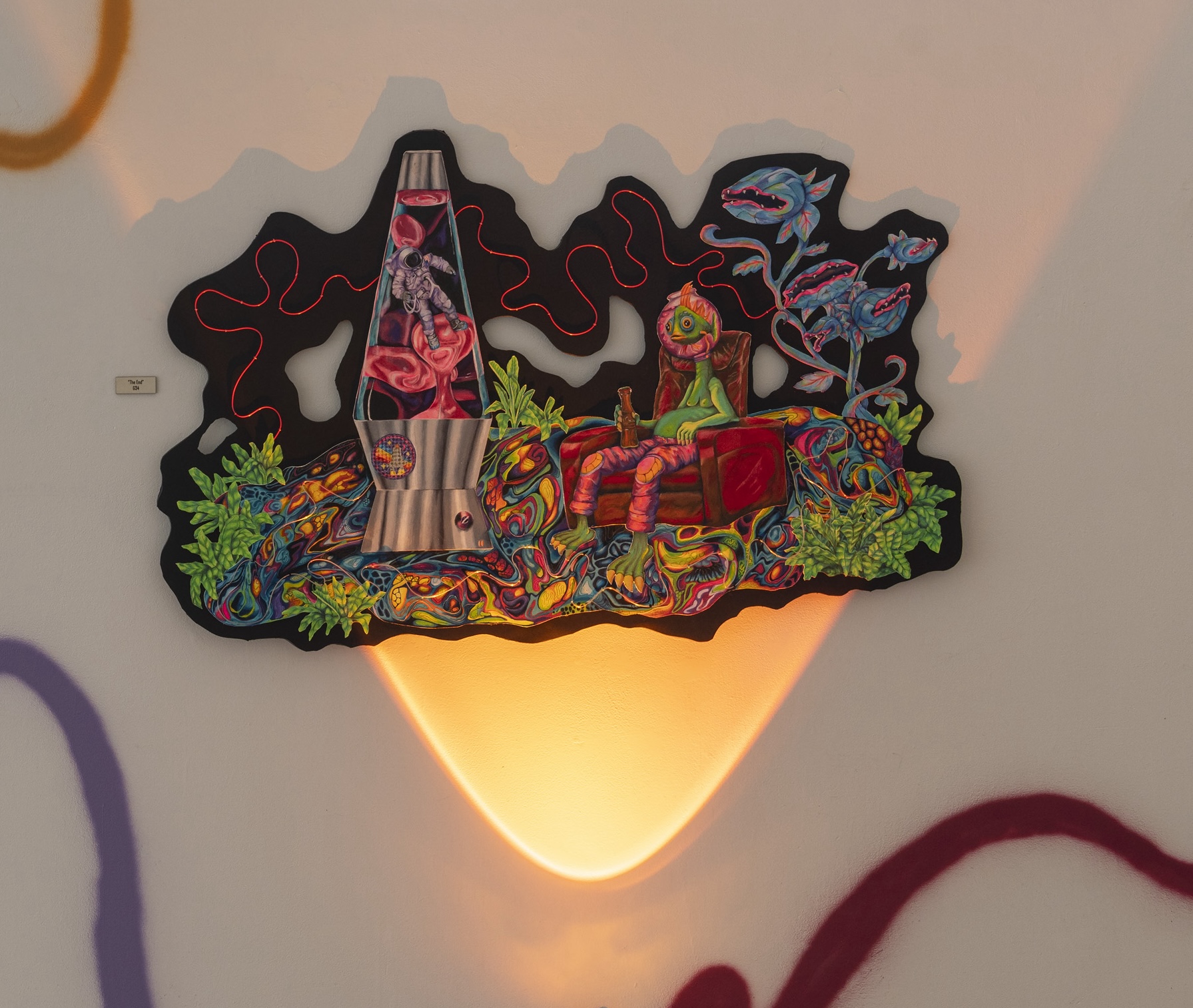 A vibrant and surreal wall-mounted artwork with a black organic-shaped background. It features a lava lamp containing an astronaut, a green humanoid figure with an oversized single eye sitting in a red chair, surrounded by colorful, abstract plants and shark-like forms. The piece is illuminated by warm light, casting a heart-shaped glow below.