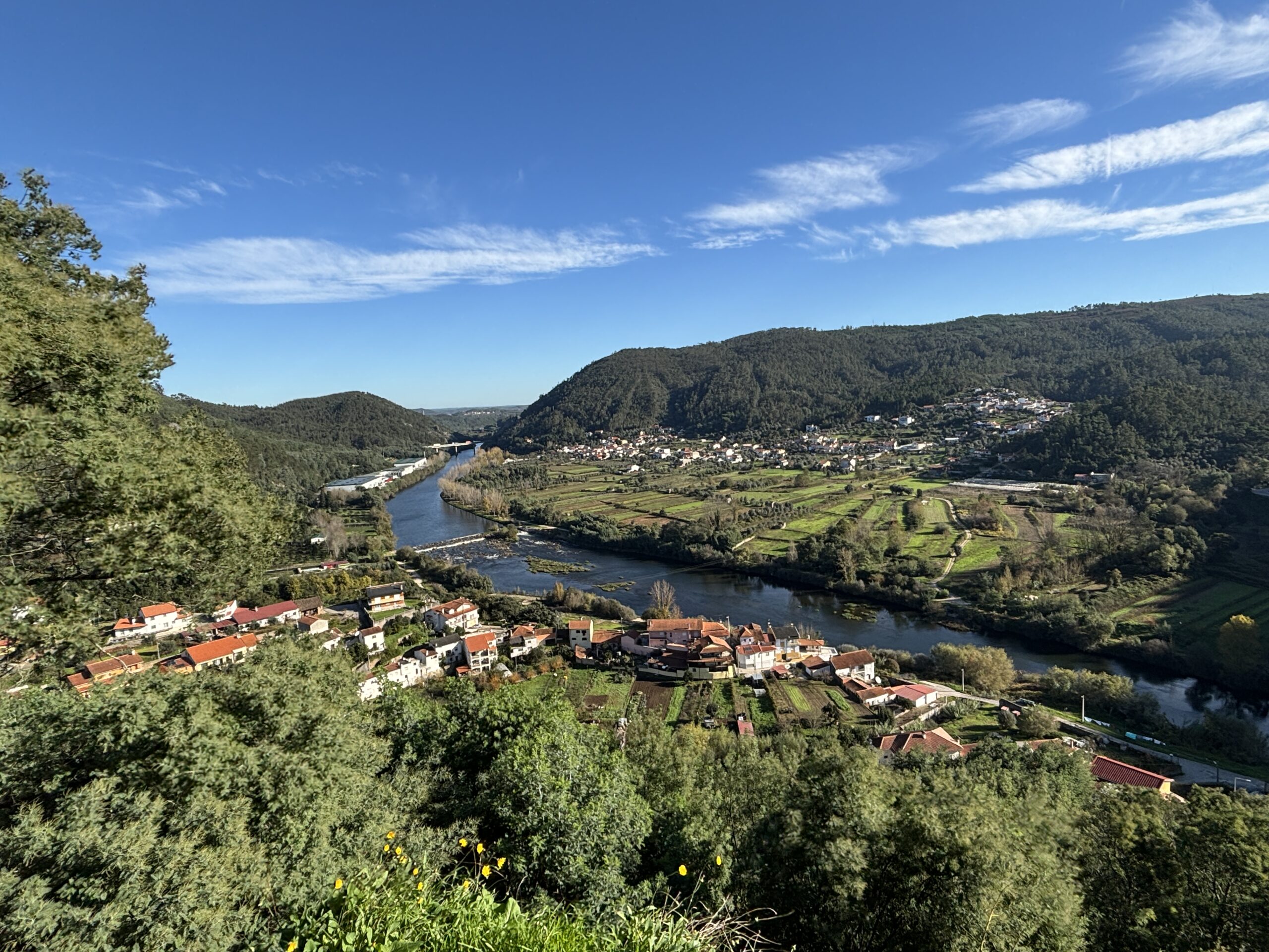 A panoramic view of a lush, green valley with the Mondego River winding through it. The landscape features a mix of small villages with red-roofed houses, agricultural fields, and dense forested hills. A blue bridge spans the river in the distance, connecting different parts of the valley. The sky is bright blue with wispy white clouds, and trees in the foreground frame the scenic countryside.