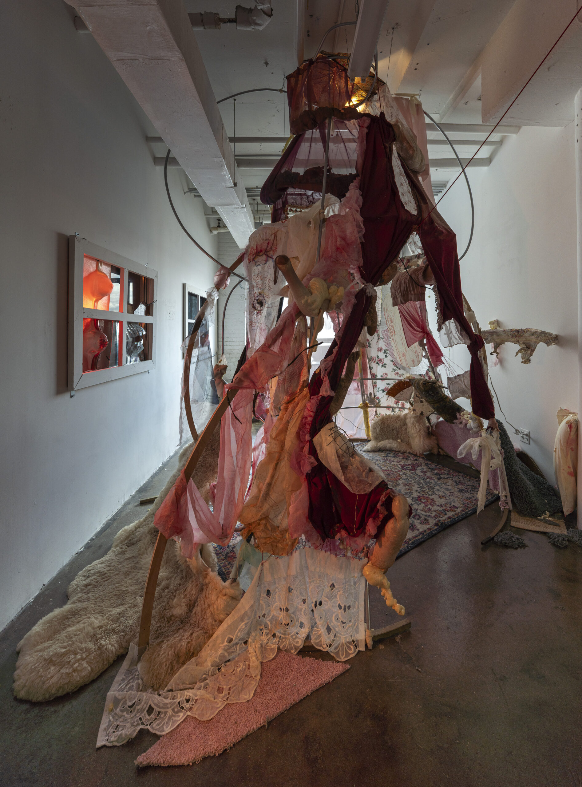 A mixed-media installation in a gallery space, featuring an elaborate sculptural structure composed of fabric, lace, and organic materials. The piece incorporates deep reds, pinks, and whites, draped and layered to create a tent-like form. Various textures, including fur rugs and patterned textiles, cover the floor. The setting includes soft lighting, and a mirror on the left wall reflects an illuminated red mannequin, adding depth and intrigue to the scene.