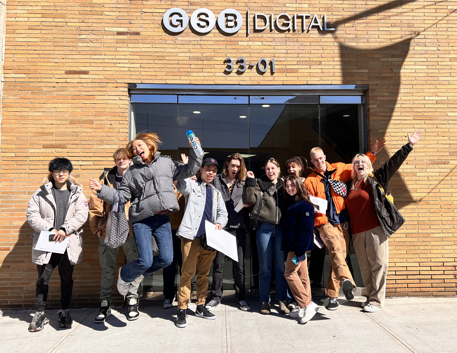 A group of ten young adults stands excitedly in front of a brick building with a sign that reads 